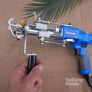 May include: A blue and grey electric tufting gun with a metal needle mechanism and a black handle. The device features a label that reads TuftingGuide and includes a small yellow component near the needle. The tool is designed for textile work.