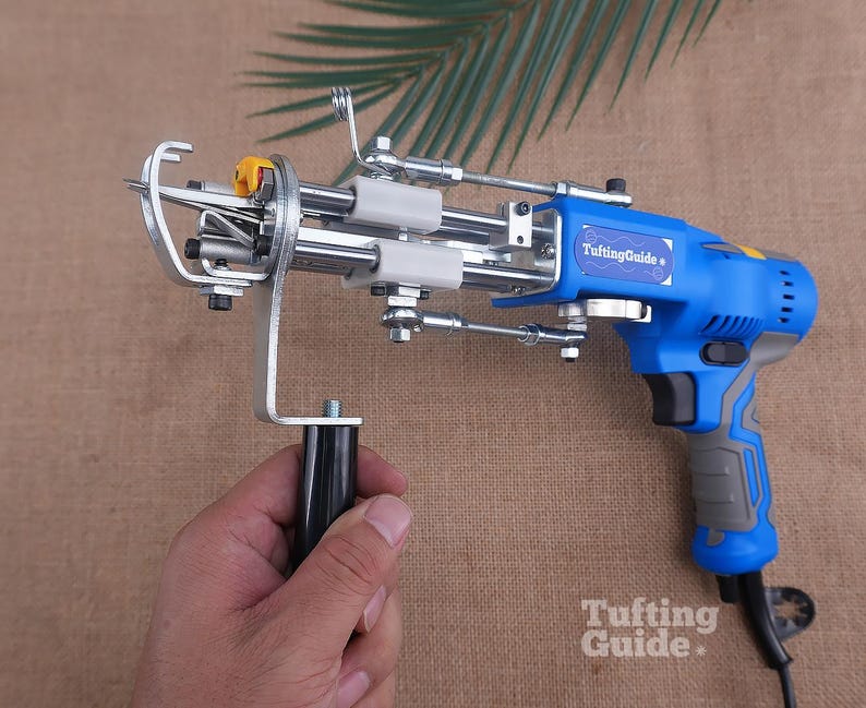 May include: A blue and grey electric tufting gun with a metal needle mechanism and a black handle. The device features a label that reads TuftingGuide and includes a small yellow component near the needle. The tool is designed for textile work.