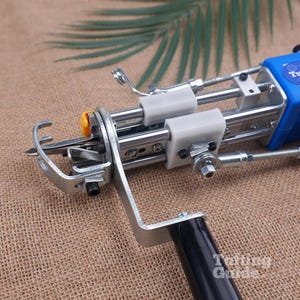 May include: A blue and silver tufting gun featuring a metal frame, a black handle, and white plastic components. The device has a needle mechanism at the front and a blue casing labelled TuftingGun. The tool is designed for textile crafting.