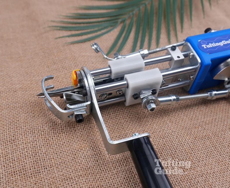 May include: A blue and silver tufting gun featuring a metal frame, a black handle, and white plastic components. The device has a needle mechanism at the front and a blue casing labelled TuftingGun. The tool is designed for textile crafting.