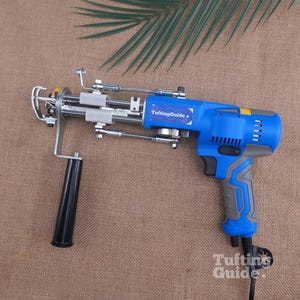 May include: A blue and grey electric tufting gun featuring a black handle, a metal needle mechanism, and a label that reads TuftingGuide. The power tool has a trigger, ventilation slots, and a power cord extending from the bottom of the grip.