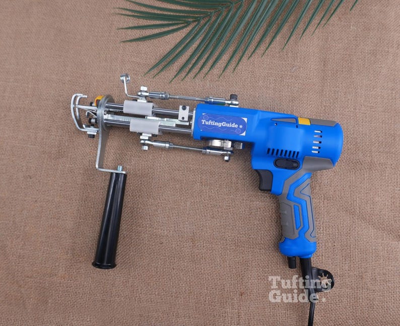 May include: A blue and grey electric tufting gun featuring a black handle, a metal needle mechanism, and a label that reads TuftingGuide. The power tool has a trigger, ventilation slots, and a power cord extending from the bottom of the grip.