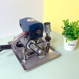 May include: A blue and silver electric machine with a black hose and a silver metal base. The machine has a belt and gears visible. The machine is sitting on a light green surface.