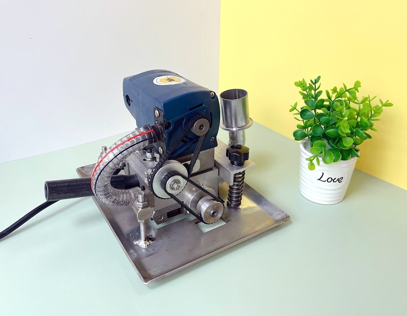 May include: A blue and silver electric machine with a black hose and a silver metal base. The machine has a belt and gears visible. The machine is sitting on a light green surface.