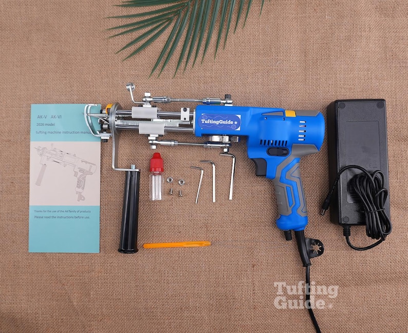 May include: A blue and grey electric tufting machine with a metal needle mechanism, a black handle, a power adapter with a cord, a small oil bottle, three L-shaped hex keys, a yellow tool, and an instruction manual labelled AK-V AK-VI 2026 model.