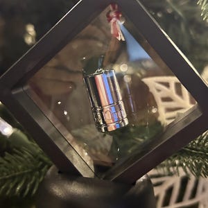 May include: A silver-coloured metal ornament shaped like a small cylinder with a black frame and a clear glass front. The ornament is hanging from a Christmas tree.