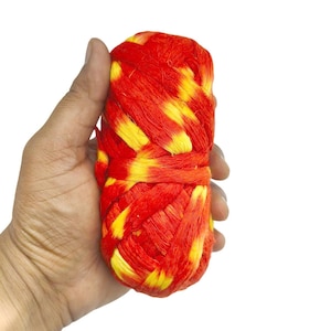 Puede incluir: Un haz cilíndrico de hilos rojos y amarillos trenzados, sostenido en una mano. Los hilos están trenzados, creando una superficie texturizada. El fondo es blanco.