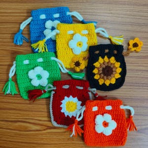 Könnte beinhalten: Eine Sammlung von sechs kleinen Häkelbeuteln mit Kordelzug in Blau, Gelb, Grün, Schwarz, Rot und Orange. Jeder Beutel ist mit einer auffälligen Häkelblumen-Applikation auf der Vorderseite und passenden Garnquasten an den Kordelenden versehen.