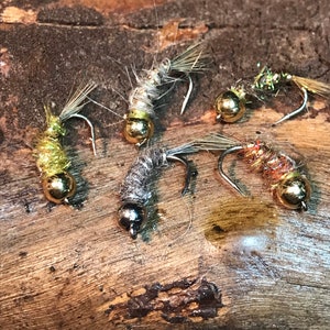 May include: Five different fly fishing lures with gold and silver beads. The lures are made with feathers and thread and are arranged on a wooden surface.