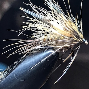 May include: A fishing fly lure featuring a gold-toned body wrapped in fine wire, adorned with long, wispy brown, tan, and black hackle feathers extending outward from the head, secured by a small metal hook at the front against a dark background.