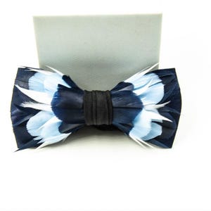 May include: A navy blue feather bow tie with light blue and white accents. The bow tie has a black center band and is set against a white background. This formal accessory is suitable for special occasions.