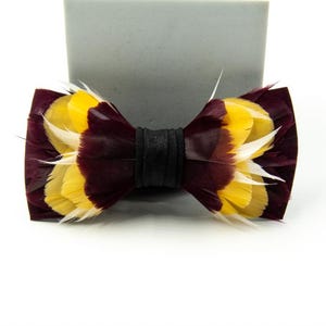 May include: A handmade bow tie featuring a striking feather design. The bow tie showcases a combination of burgundy, yellow, and white feathers, with a black band in the center. This accessory is perfect for adding a touch of elegance to any outfit.