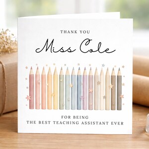 May include: A white card with the text "Thank You Miss Cole" and "For Being The Best Teaching Assistant Ever." The card features pastel-coloured pencils and star accents. A wrapped gift and rolls of paper are in the background.