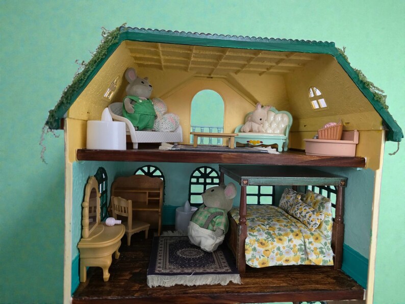K&ouml;nnte beinhalten: Ein mehrst&ouml;ckiges Puppenhaus mit gr&uuml;nem Dach und gelben W&auml;nden. Das Haus ist mit Miniaturm&ouml;beln eingerichtet und enth&auml;lt M&auml;usefiguren. Das Puppenhaus hat ein Bett, ein Sofa, einen Schminktisch und anderes Zubeh&ouml;r. Das Haus ist etwa 30 cm hoch.