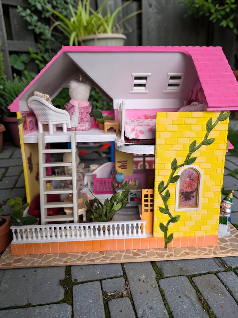 May include: A colorful dollhouse with a pink roof, yellow brick walls, and a white balcony. The dollhouse has multiple levels, a ladder, and a small figure on the balcony. A green vine climbs up the side.