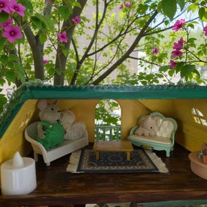 K&ouml;nnte beinhalten: Miniatur-Puppenhaus-Interieur mit gr&uuml;nem Dach und gelben W&auml;nden. Im Inneren befinden sich ein kleines Sofa mit einer Mausfigur in gr&uuml;nen Overalls, ein kleiner Tisch und ein Teppich. Der Hintergrund zeigt rosa Bl&uuml;ten und gr&uuml;ne Bl&auml;tter.
