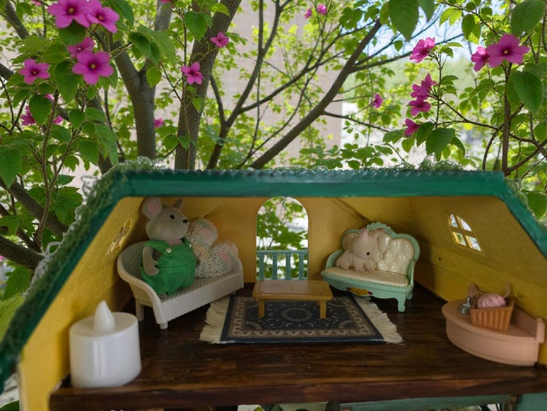 K&ouml;nnte beinhalten: Miniatur-Puppenhaus-Interieur mit gr&uuml;nem Dach und gelben W&auml;nden. Im Inneren befinden sich ein kleines Sofa mit einer Mausfigur in gr&uuml;nen Overalls, ein kleiner Tisch und ein Teppich. Der Hintergrund zeigt rosa Bl&uuml;ten und gr&uuml;ne Bl&auml;tter.