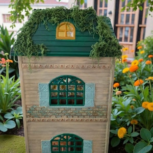 K&ouml;nnte beinhalten: Ein Miniatur-Puppenhaus aus Holz mit einem gr&uuml;nen Dach, das mit Gr&uuml;n bedeckt ist. Das Haus hat zwei gew&ouml;lbte Fenster mit gr&uuml;nen Rahmen und blauen Fensterl&auml;den. Die W&auml;nde sind hellbeige mit Ziegeldetails.