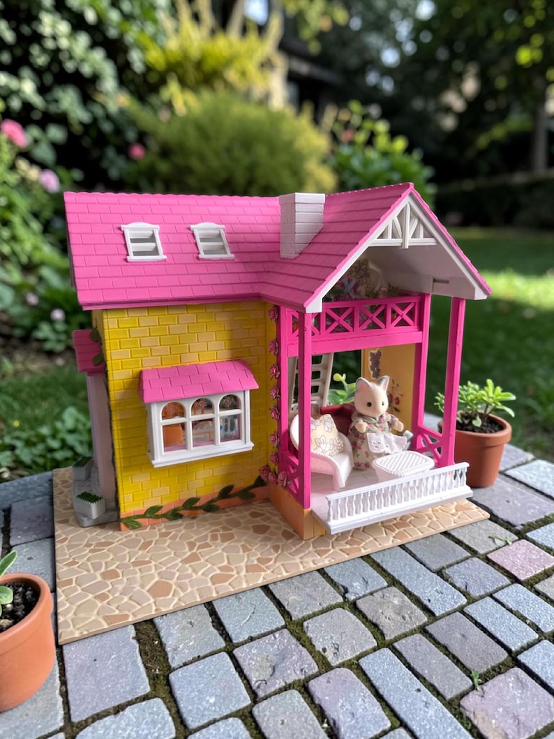 May include: A miniature dollhouse with a pink roof, yellow brick walls, and a porch. The porch has a white railing and a small table. A doll dressed in a floral print dress is seated on a white bench. The dollhouse is set on a stone patio.