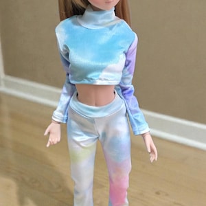 May include: A doll with long blonde hair and blue eyes wearing a tie-dye outfit. The outfit includes a cropped, long-sleeved top and flared pants in shades of blue, purple, and pink. White sneakers complete the look.