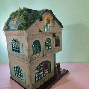 K&ouml;nnte beinhalten: Ein detailliertes Puppenhaus mit einem gr&uuml;nen Dach, das mit k&uuml;nstlichem Laub bedeckt ist. Das Haus hat mehrere Fenster mit t&uuml;rkisfarbenen Rahmen und einen kleinen Balkon. Eine kleine Figur ist auf dem Balkon zu sehen. Das Haus ist in neutralen T&ouml;nen mit dekorativen Zierleisten bemalt.