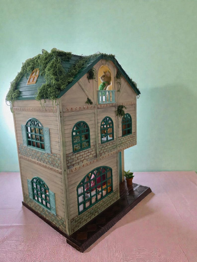 K&ouml;nnte beinhalten: Ein detailliertes Puppenhaus mit einem gr&uuml;nen Dach, das mit k&uuml;nstlichem Laub bedeckt ist. Das Haus hat mehrere Fenster mit t&uuml;rkisfarbenen Rahmen und einen kleinen Balkon. Eine kleine Figur ist auf dem Balkon zu sehen. Das Haus ist in neutralen T&ouml;nen mit dekorativen Zierleisten bemalt.