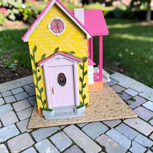 May include: A miniature yellow house with a pink roof, door, and porch. The house has a round window and is decorated with green vines. The house sits on a stone patio.