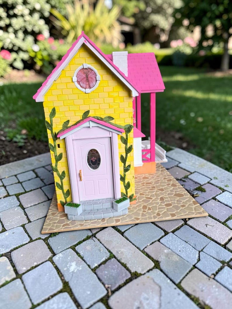 May include: A miniature yellow house with a pink roof, door, and porch. The house has a round window and is decorated with green vines. The house sits on a stone patio.