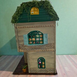 K&ouml;nnte beinhalten: Ein Miniatur-Zweifamilienhaus mit einem gr&uuml;nen, mit Moos bedeckten Dach. Das Haus hat eine hellbraune Holzfassade mit Ziegelakzenten, blaue Fensterl&auml;den und gew&ouml;lbte Fenster. Eine kleine Topfpflanze steht auf dem Sockel.