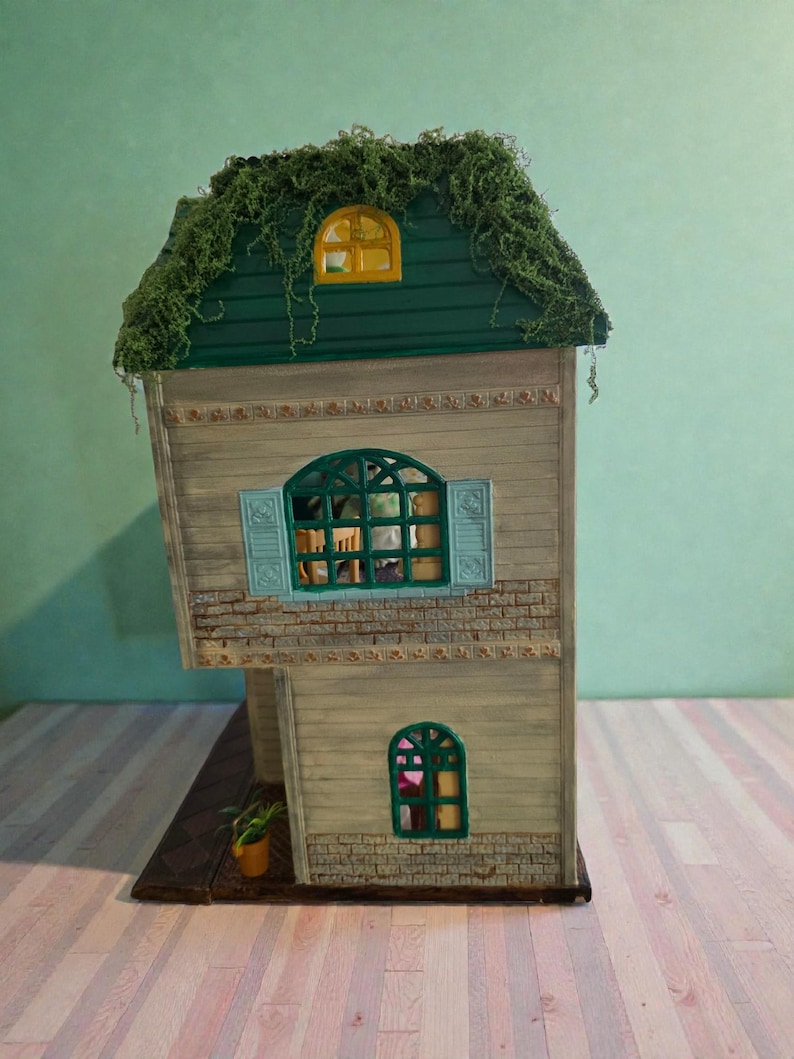 K&ouml;nnte beinhalten: Ein Miniatur-Zweifamilienhaus mit einem gr&uuml;nen, mit Moos bedeckten Dach. Das Haus hat eine hellbraune Holzfassade mit Ziegelakzenten, blaue Fensterl&auml;den und gew&ouml;lbte Fenster. Eine kleine Topfpflanze steht auf dem Sockel.
