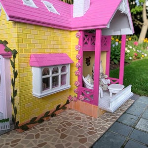 May include: A miniature dollhouse with a yellow brick exterior, a pink roof, and a white chimney. The porch has pink columns and a white railing. Pink roses decorate the columns. A small table and chair set are on the porch.