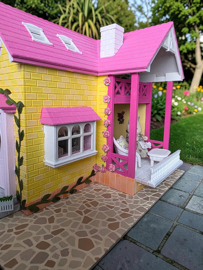 May include: A miniature dollhouse with a yellow brick exterior, a pink roof, and a white chimney. The porch has pink columns and a white railing. Pink roses decorate the columns. A small table and chair set are on the porch.