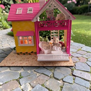 May include: A miniature dollhouse with a yellow exterior and a pink roof. The house features a balcony with a white railing and a small doll sitting on a white chair. The house is set on a stone patio.