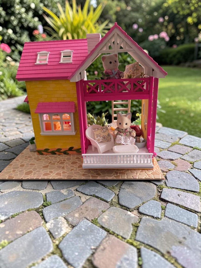 May include: A miniature dollhouse with a yellow exterior and a pink roof. The house features a balcony with a white railing and a small doll sitting on a white chair. The house is set on a stone patio.