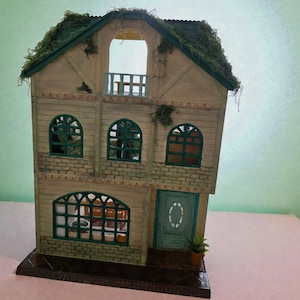 K&ouml;nnte beinhalten: Ein Miniatur-Zweifamilienhaus mit gr&uuml;nem Dach und Zierleisten. Das Haus hat gew&ouml;lbte Fenster, einen Balkon und eine t&uuml;rkisfarbene T&uuml;r. Die Fassade ist in Beige- und Gr&uuml;nt&ouml;nen gehalten, mit einer kleinen Topfpflanze in der N&auml;he der T&uuml;r.