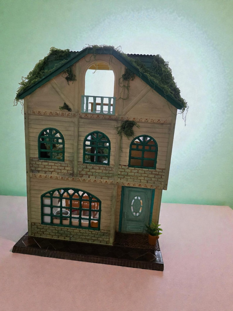 K&ouml;nnte beinhalten: Ein Miniatur-Zweifamilienhaus mit gr&uuml;nem Dach und Zierleisten. Das Haus hat gew&ouml;lbte Fenster, einen Balkon und eine t&uuml;rkisfarbene T&uuml;r. Die Fassade ist in Beige- und Gr&uuml;nt&ouml;nen gehalten, mit einer kleinen Topfpflanze in der N&auml;he der T&uuml;r.