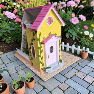May include: A miniature yellow house with a pink roof and door, adorned with green vines. The house has a small white fence and is set on a brick patio, with small potted plants nearby. The background features a garden with pink flowers.