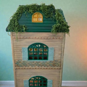 K&ouml;nnte beinhalten: Ein Holzpuppenhaus mit gr&uuml;nem Dach und kleinem gelben Fenster. Das Haus hat zwei gr&uuml;ne Bogenfenster mit blauen Fensterl&auml;den. Die Fassade ist in hellen Holzt&ouml;nen mit Ziegelakzenten gehalten.