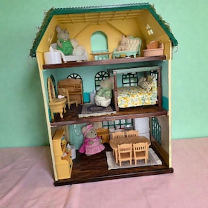 K&ouml;nnte beinhalten: Ein dreist&ouml;ckiges Puppenhaus mit gr&uuml;nem Dach und gelben W&auml;nden. Das Puppenhaus ist mit Miniaturm&ouml;beln eingerichtet und enth&auml;lt M&auml;usefiguren in verschiedenen R&auml;umen. Zu den R&auml;umen geh&ouml;ren ein Schlafzimmer, ein Wohnzimmer und ein Esszimmer.