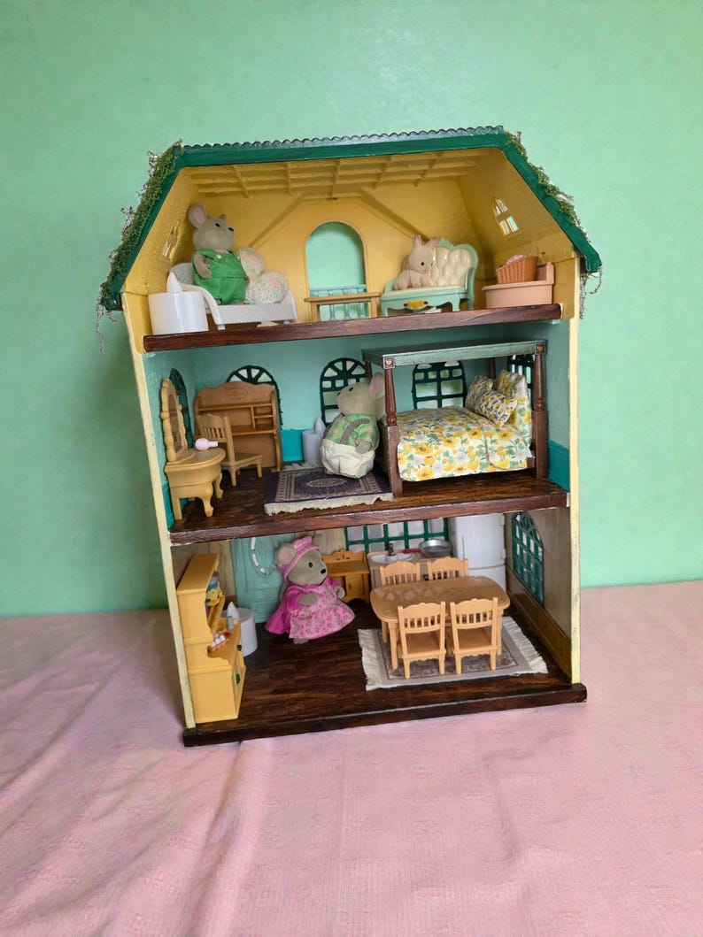 K&ouml;nnte beinhalten: Ein dreist&ouml;ckiges Puppenhaus mit gr&uuml;nem Dach und gelben W&auml;nden. Das Puppenhaus ist mit Miniaturm&ouml;beln eingerichtet und enth&auml;lt M&auml;usefiguren in verschiedenen R&auml;umen. Zu den R&auml;umen geh&ouml;ren ein Schlafzimmer, ein Wohnzimmer und ein Esszimmer.