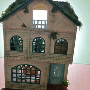 K&ouml;nnte beinhalten: Ein Miniatur-Puppenhaus mit hellbrauner Fassade, gr&uuml;nen Zierleisten und einem gr&uuml;nen Dach, das mit k&uuml;nstlichem Gr&uuml;n bedeckt ist. Das Haus verf&uuml;gt &uuml;ber gew&ouml;lbte Fenster, einen Balkon und eine gr&uuml;ne Eingangst&uuml;r. Eine kleine Topfpflanze steht an der T&uuml;r.