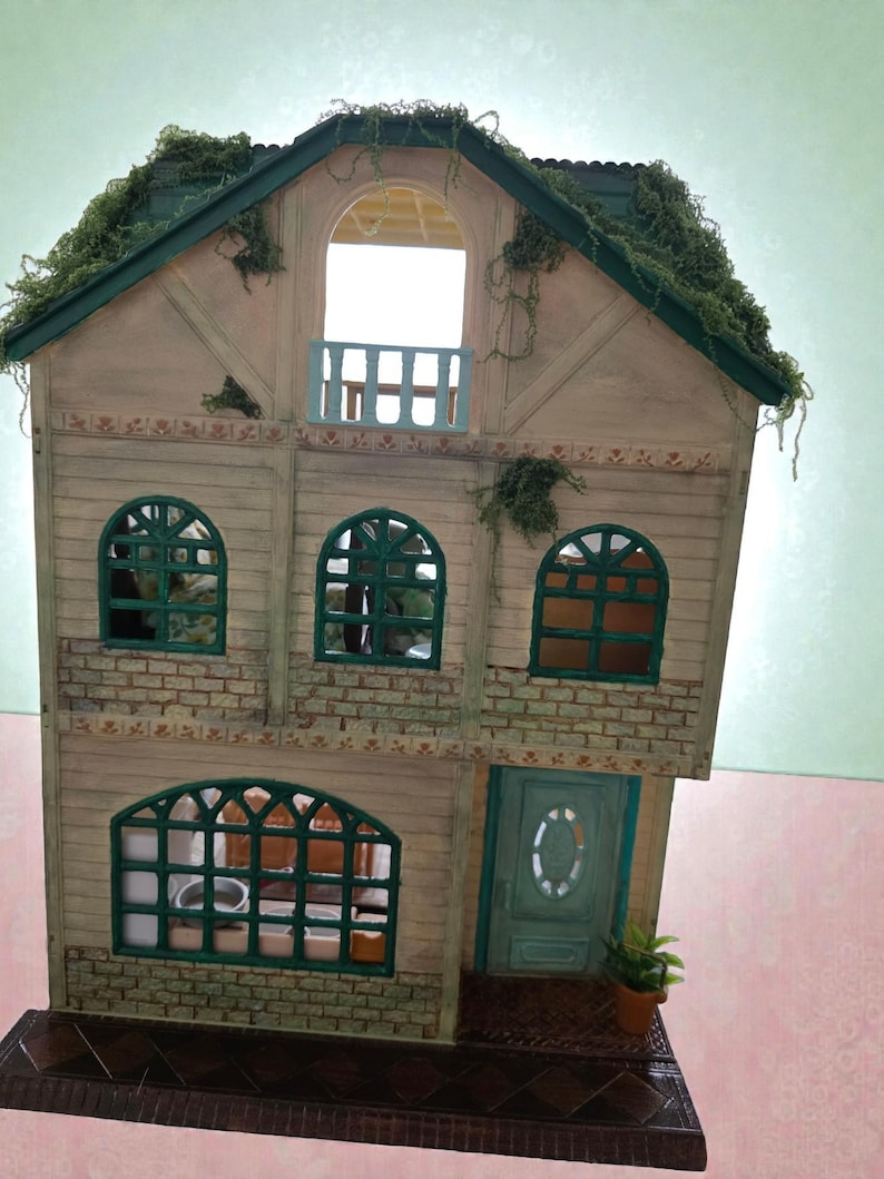 K&ouml;nnte beinhalten: Ein Miniatur-Puppenhaus mit hellbrauner Fassade, gr&uuml;nen Zierleisten und einem gr&uuml;nen Dach, das mit k&uuml;nstlichem Gr&uuml;n bedeckt ist. Das Haus verf&uuml;gt &uuml;ber gew&ouml;lbte Fenster, einen Balkon und eine gr&uuml;ne Eingangst&uuml;r. Eine kleine Topfpflanze steht an der T&uuml;r.