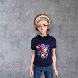 May include: A doll dressed in a navy blue short-sleeved top featuring a vibrant leopard design, paired with blue jeans and a brown and white knitted cap. The doll has blonde hair and blue eyes. The top has a black collar.
