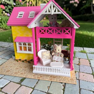 May include: A miniature dollhouse with a yellow exterior and a pink roof and balcony. The house features white trim, windows, and a small porch with a white railing. Two small animal figurines are on the balcony and porch. The house is set on a stone patio.