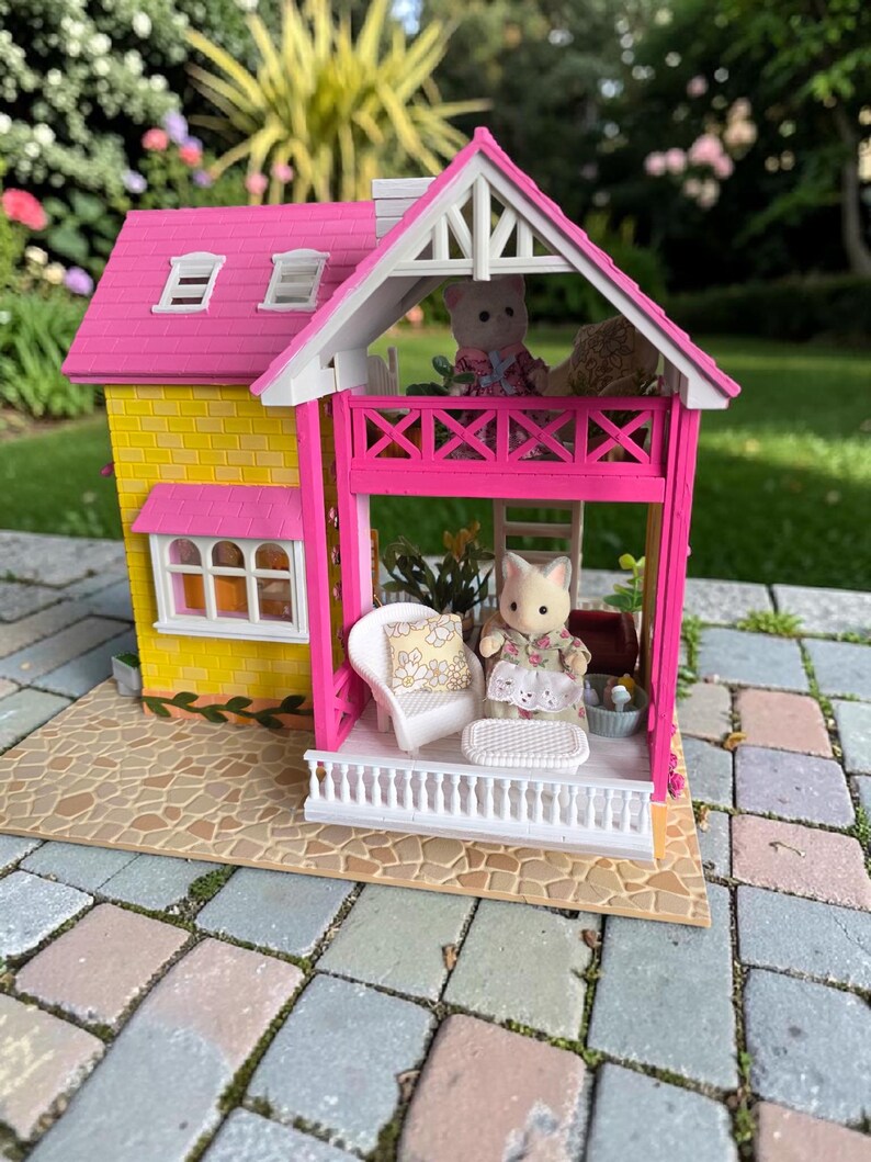 May include: A miniature dollhouse with a yellow exterior and a pink roof and balcony. The house features white trim, windows, and a small porch with a white railing. Two small animal figurines are on the balcony and porch. The house is set on a stone patio.