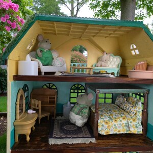 K&ouml;nnte beinhalten: Ein detailliertes Puppenhaus mit mehreren Zimmern, eingerichtet mit Miniaturm&ouml;beln und Figuren. Das Haus hat eine gelbe Fassade mit gr&uuml;nen Zierleisten und einem gr&uuml;nen Dach. Die Zimmer sind mit Miniaturm&ouml;beln eingerichtet, darunter ein Bett, ein Sofa und ein Schminktisch. Das Haus befindet sich im Freien.