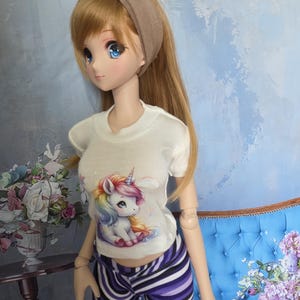 May include: A doll wearing a white t-shirt with a colorful unicorn graphic, paired with purple and white striped pants. The doll has long blonde hair, blue eyes, and a brown headband. The background features a blue wall and a blue couch.