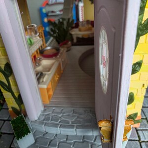 May include: A miniature dollhouse with an open door revealing a kitchen and living area. The exterior features yellow brick, pink trim, and a small stone staircase. A small green plant is visible on the left side of the entrance.