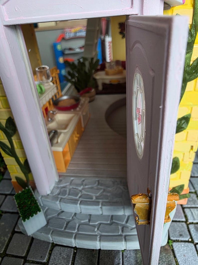 May include: A miniature dollhouse with an open door revealing a kitchen and living area. The exterior features yellow brick, pink trim, and a small stone staircase. A small green plant is visible on the left side of the entrance.