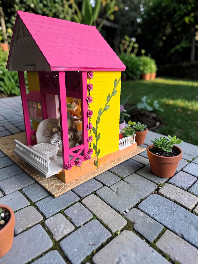 May include: A miniature dollhouse with a bright pink roof and yellow walls. The house has a porch with a white railing and is decorated with pink roses and green vines. Small potted plants sit nearby on a brick patio.